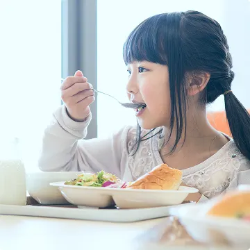 明るい室内のテーブルで、スープやサラダ、パン、牛乳などが並んだトレイの前に、スプーンで食事をする女の子の横顔。