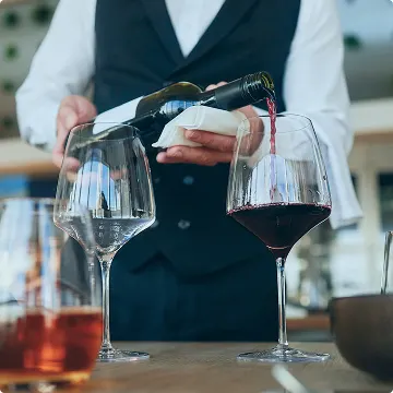 ベストを着たウェイターが、テーブルに置かれたワイングラスに赤ワインを丁寧に注いでいます。